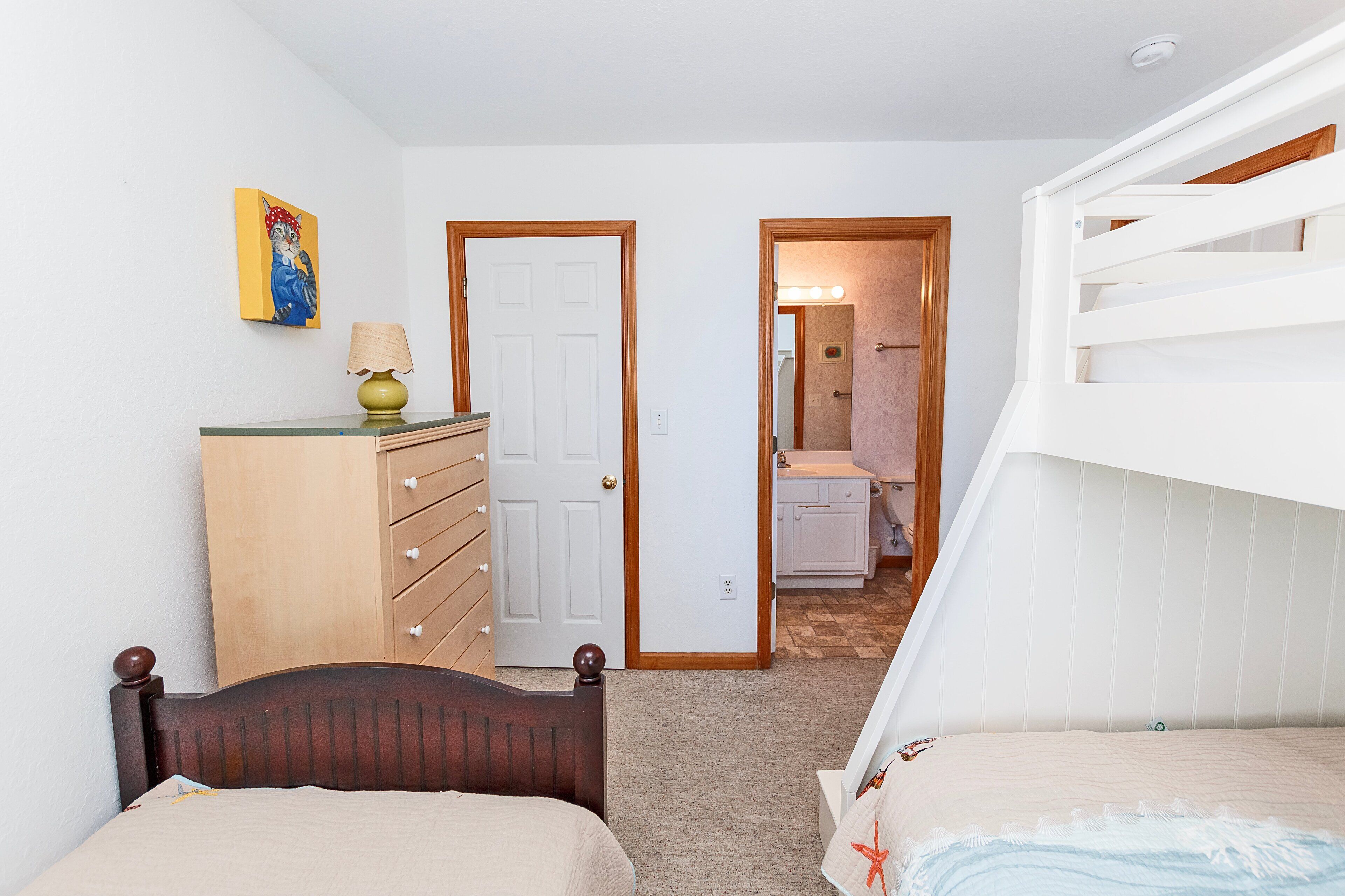 Une chambre avec un lit superposé, une commode en bois, une porte donnant sur une salle de bain et un tableau accroché au mur.
