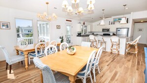 A spacious dining area with a wooden table and white wicker chairs, a kitchen with white cabinets and modern appliances, and large windows offering a view of the outdoors.