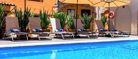 A poolside area with lounge chairs and umbrellas, a clear blue pool, and a building with a stone facade.