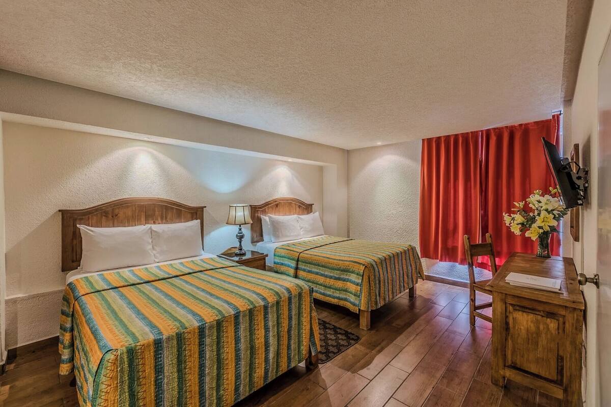 A hotel room with two beds, a wooden desk, a chair, and a red curtain.