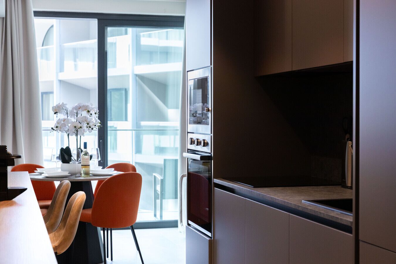 Une cuisine moderne dotée d’appareils électroménagers en acier inoxydable, un coin repas avec des chaises orange et une vue sur un immeuble à travers de grandes fenêtres.