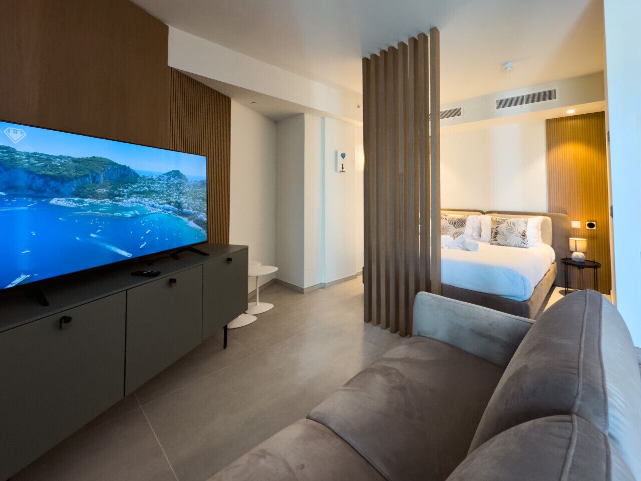 A modern hotel room with a large flat-screen TV, a sofa, a bed, and a small side table.