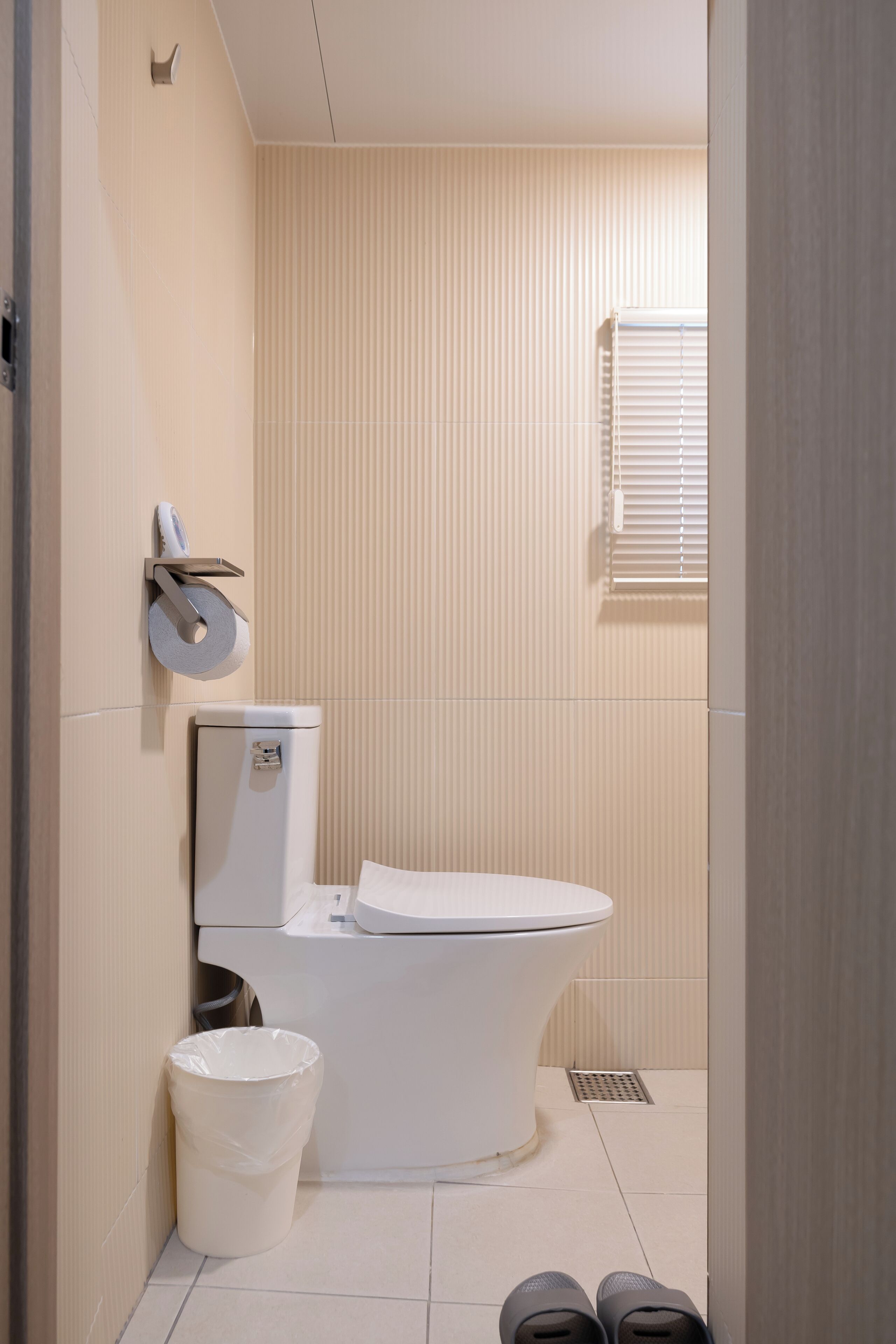 A modern bathroom with a toilet, a trash bin, and a window with blinds.