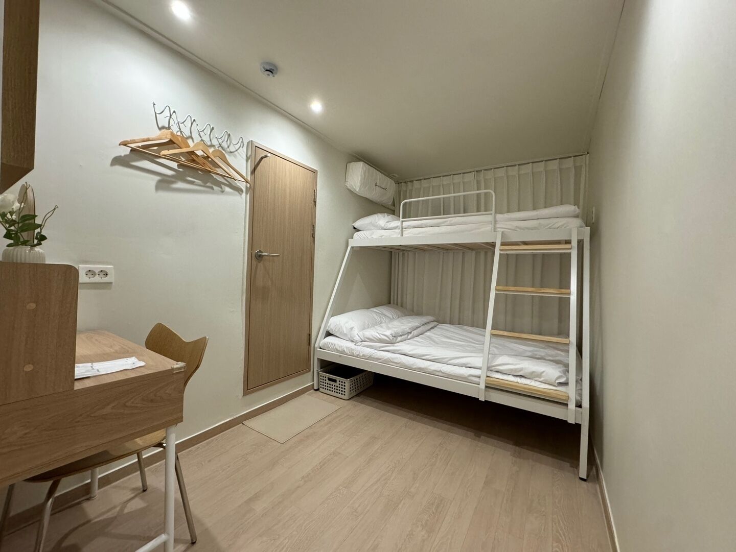 A small, well-lit room with a bunk bed, a wooden desk, and a chair. There is a door and a wall-mounted shelf with clothes.