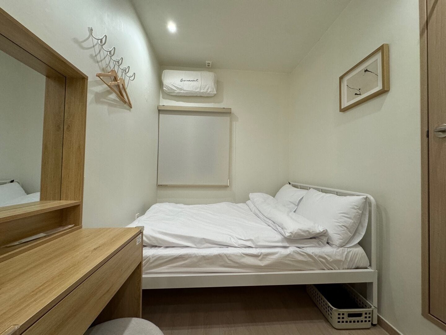 A small, neatly organized bedroom with a wooden dresser, a single bed with white bedding, a wall-mounted air conditioner, and a framed picture on the wall.