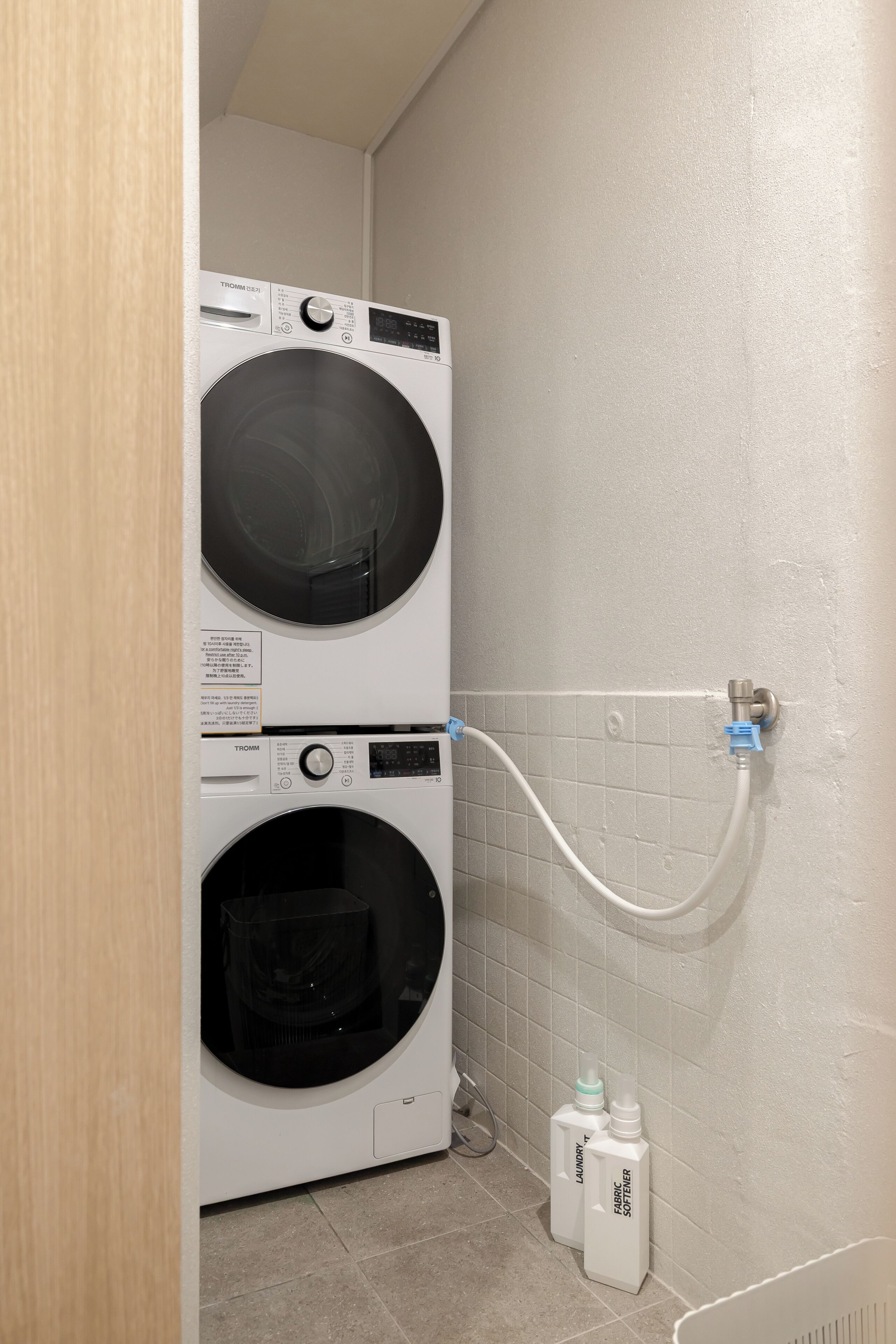 A laundry area with two stacked washing machines, a water connection, and two bottles of laundry detergent.