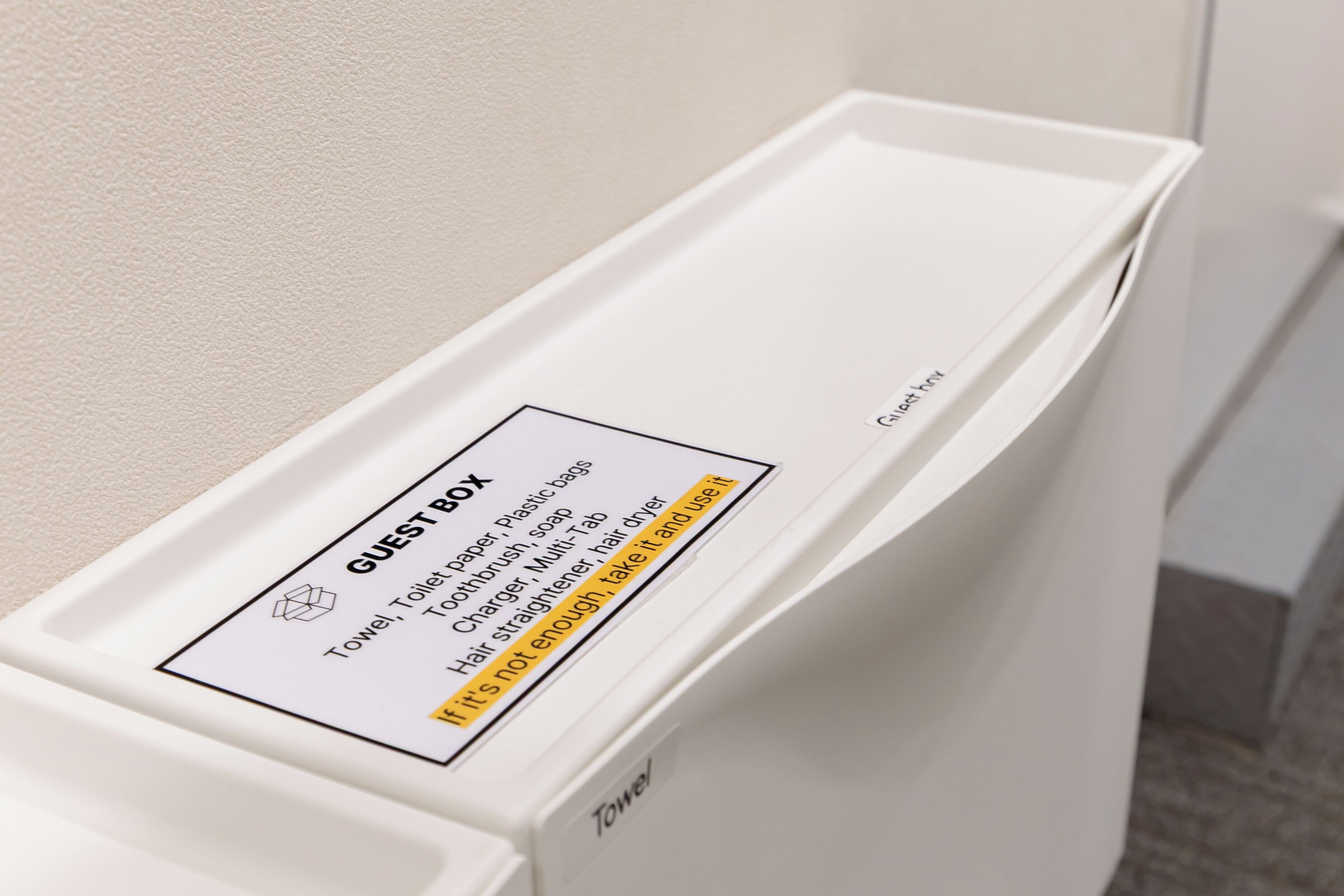 A white guest box with a label listing items like a towel, toilet paper, and a hair dryer.