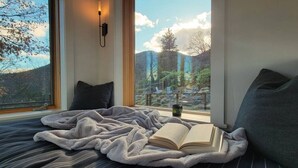A bedroom with a large window, a bed with striped bedding, a book, and a nightstand with a lamp. - Hot tub | Sauna |Firepit |Sleeps 6! (Ashland)