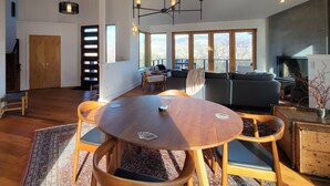 A modern living room with a large window, a fireplace, and a dining area with a round table and chairs. - Hot tub | Sauna |Firepit |Sleeps 6! (Ashland)