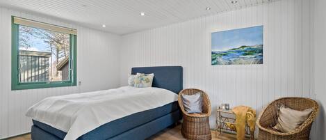 Quarto com cama, cadeiras de vime, um quadro e janela com vista para árvores.