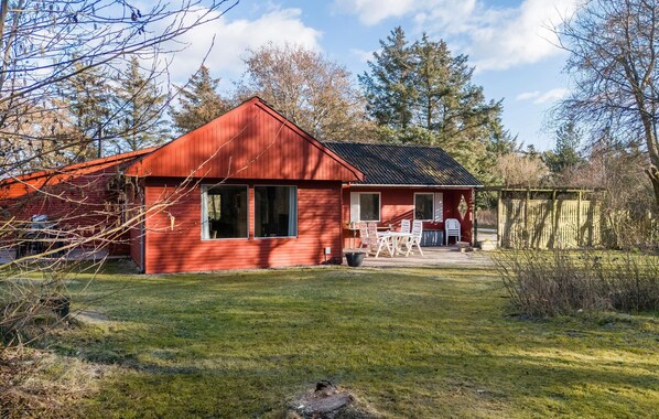 A red wooden cabin with a patio, surrounded by trees and a grassy yard.