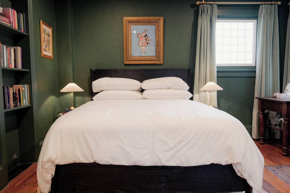 Una cama bien hecha con ropa blanca, cabecera de madera oscura y dos lámparas de noche. A la izquierda, una estantería llena de libros, y encima de la cama, un cuadro enmarcado.