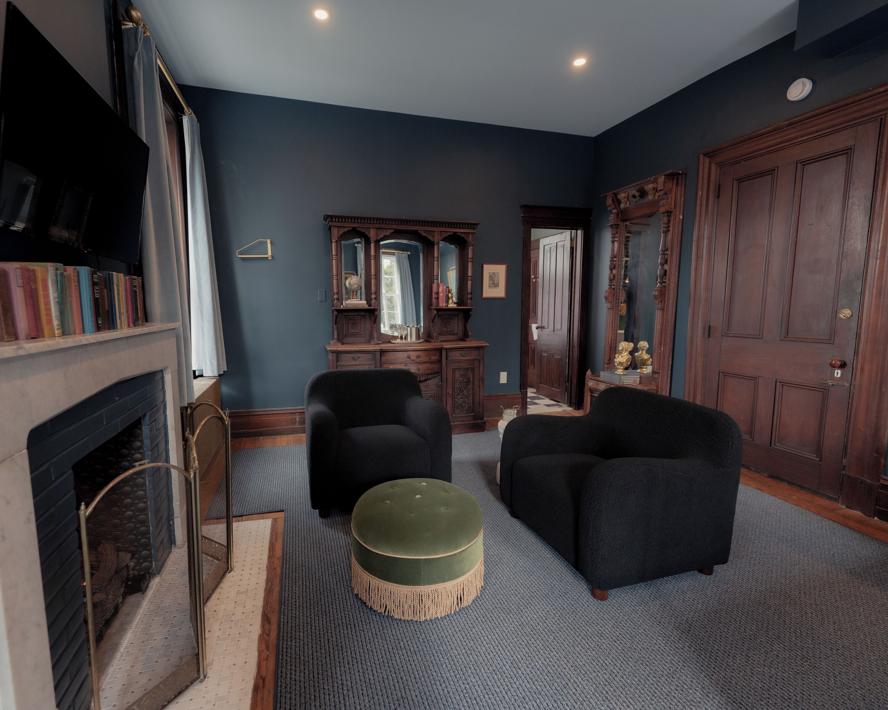 Une pièce avec une cheminée, deux fauteuils, un pouf rond, un meuble en bois avec un miroir et une grande porte en bois.