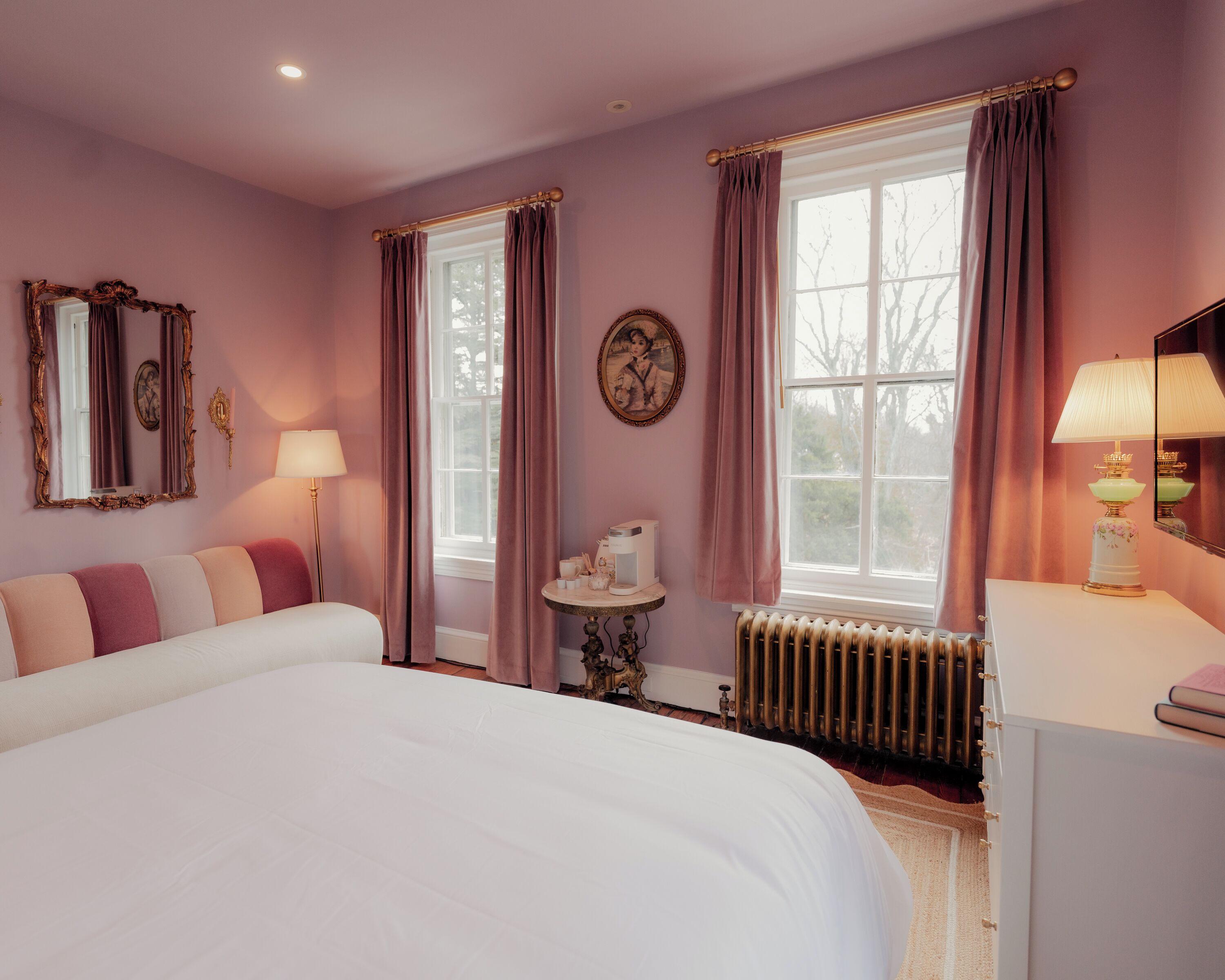 Une chambre à coucher avec un grand lit, une commode, un radiateur et deux fenêtres avec des rideaux.