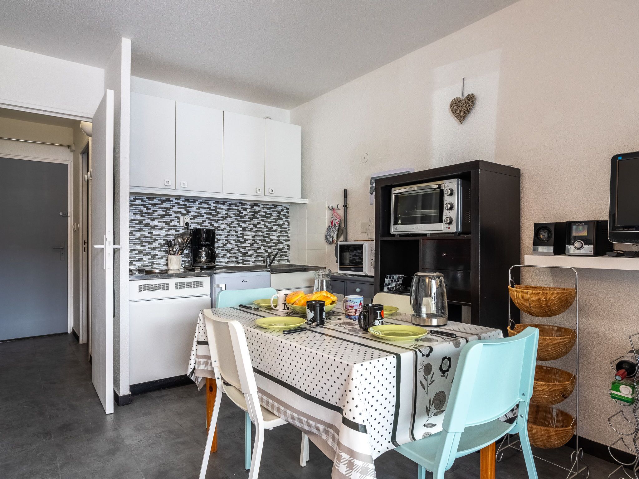 A compact kitchen with white cabinets, a coffee maker, a toaster, and a microwave. There is a dining table with chairs and a basket of fruits.