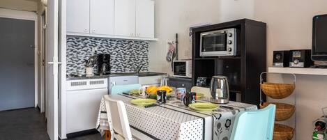 A compact kitchen with white cabinets, a coffee maker, a toaster, and a microwave. There is a dining table with chairs and a basket of fruits.