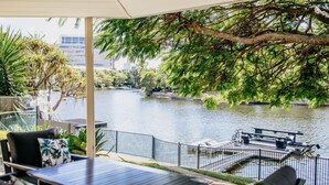 A riverside patio with outdoor seating, a boat dock, and a view of the river and cityscape. - Riverside Retreat Pool, Pontoon & Space (Bundall)