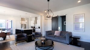 A modern living room with a grey sofa, two armchairs, a coffee table, and a kitchen in the background. - Songbird Suites - 2 Rooftops & Outdoor Cinema (Nashville)