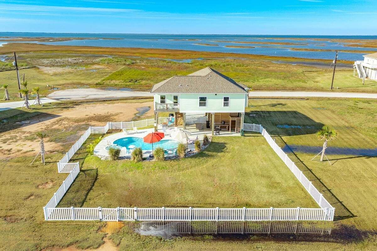 A two-story house with a swimming pool, surrounded by a white fence, located near a beach.