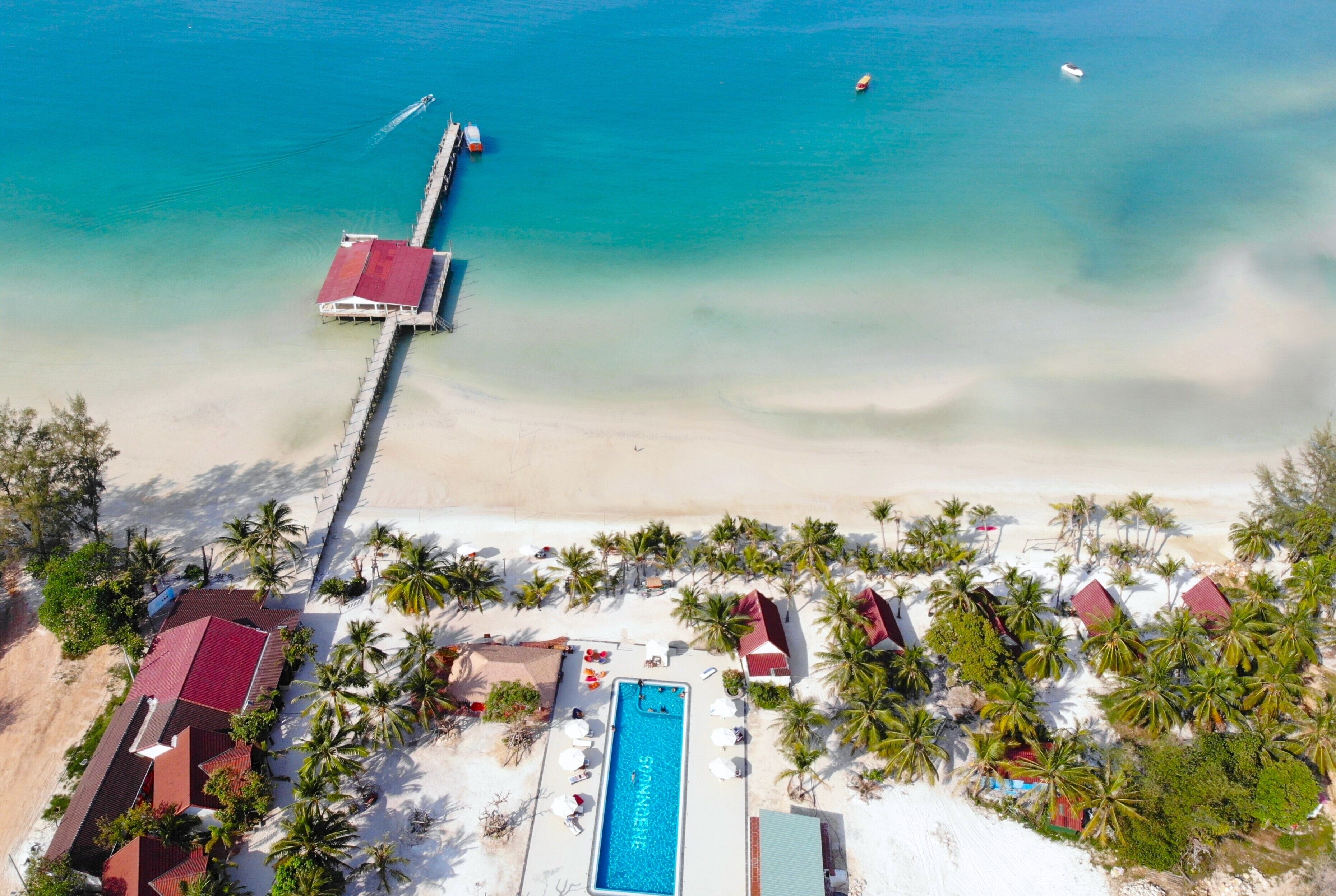 An aerial view of a beach resort with a swimming pool, palm trees, and a pier extending into the sea.