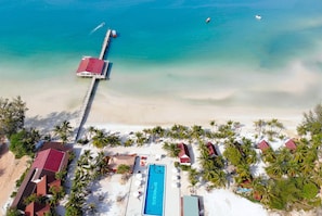 Vista aérea de un complejo turístico en la playa con piscina, palmeras y un muelle que se adentra en el mar.