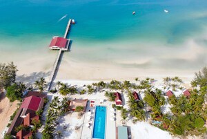 An aerial view of a beach resort with a swimming pool, palm trees, and a pier extending into the sea.