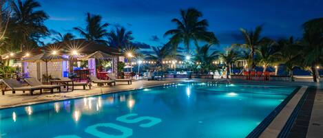 A swimming pool with lounge chairs and umbrellas, surrounded by palm trees and a night sky.