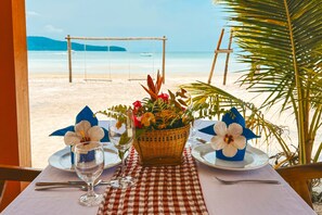 Una mesa con mantel a cuadros, un vaso de agua, una cesta de flores y vista a una playa con un columpio.