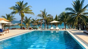 Ein Swimmingpool mit Liegestühlen und Sonnenschirmen, umgeben von Palmen und Blick aufs Meer.