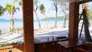 Eine Terrasse am Strand mit Backsteinpfeilern, einem Holztisch und Stühlen, mit Blick auf einen Sandstrand mit Palmen und eine klare Meerblick.