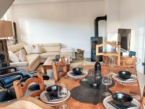 A modern living room with a sofa, coffee table, and dining table set for a meal. A wood-burning stove is visible in the background.
