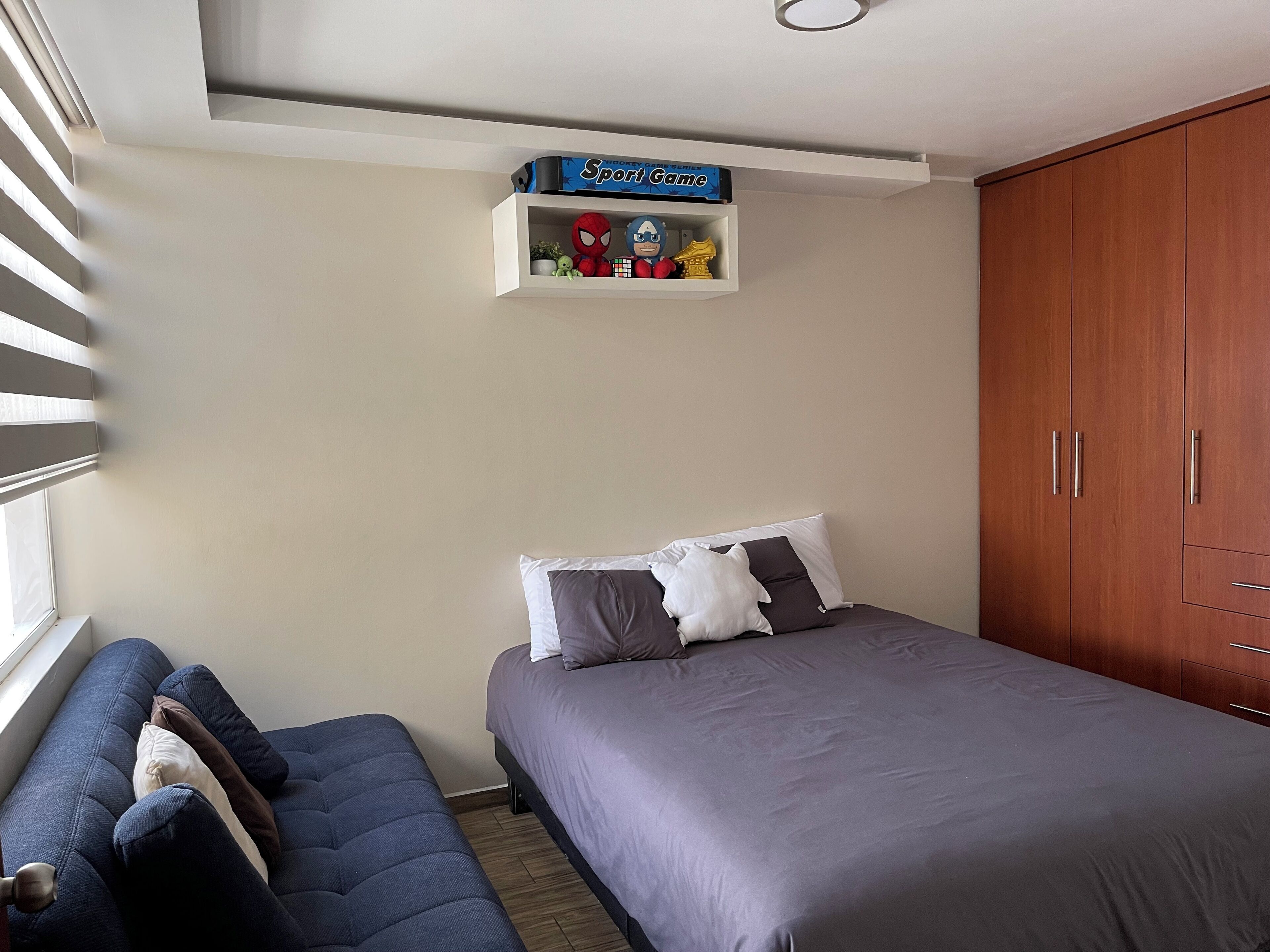 Une chambre à coucher avec un lit, une armoire, une étagère remplie de jouets et une fenêtre avec des stores.