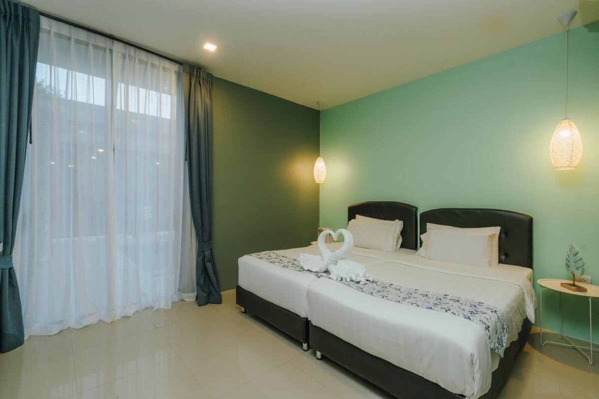 A double bed with white bedding and a heart-shaped towel arrangement, a bedside table with a lamp, and a view of the outdoors through sheer curtains.