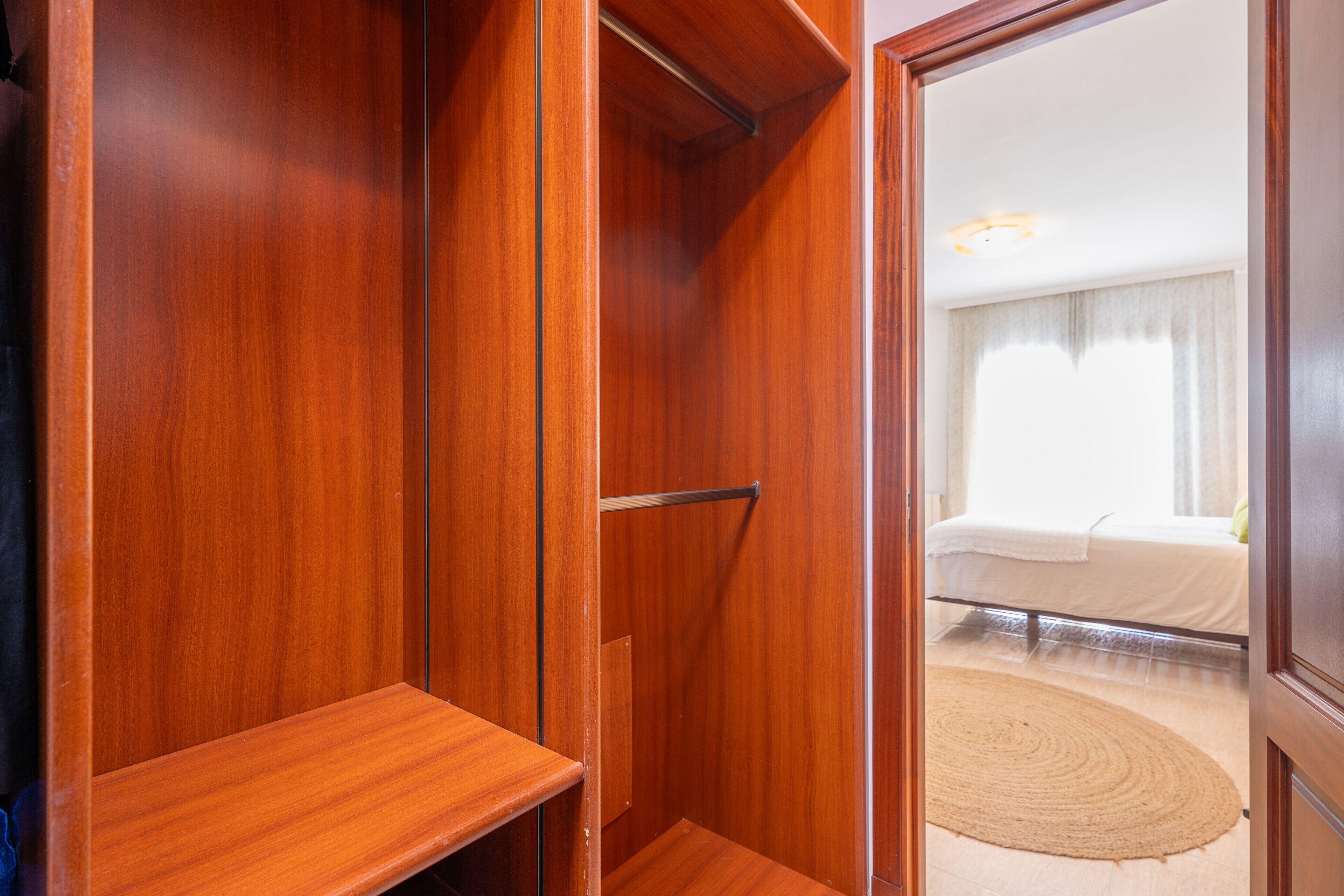 Un armario de madera con barra metálica, un estante pequeño y una puerta que da a un dormitorio con cama y alfombra redonda.