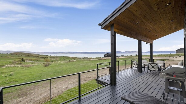 A wooden deck with outdoor seating and a barbecue grill overlooking a grassy landscape and a body of water.