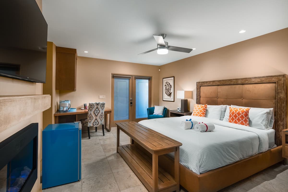 A hotel room with a bed, a desk, a chair, a small table, a ceiling fan, and a TV.