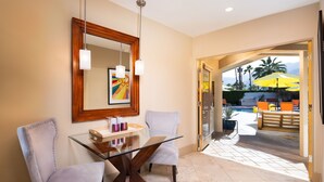 A dining area with a glass table and two chairs, a wooden-framed mirror, and a view of an outdoor pool area with umbrellas. - El Noa Noa (Palm Springs)