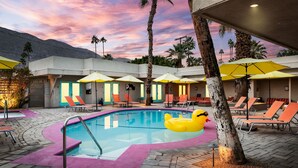 A pool area with lounge chairs and umbrellas, surrounded by palm trees and a building with a modern design. - El Noa Noa (Palm Springs)