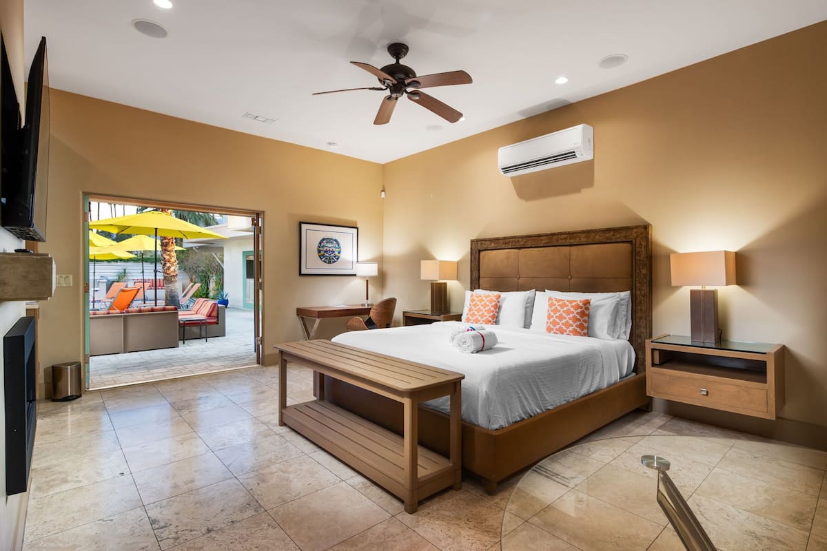 A hotel room with a bed, bedside tables, a ceiling fan, an air conditioning unit, and a view of the outdoor seating area.