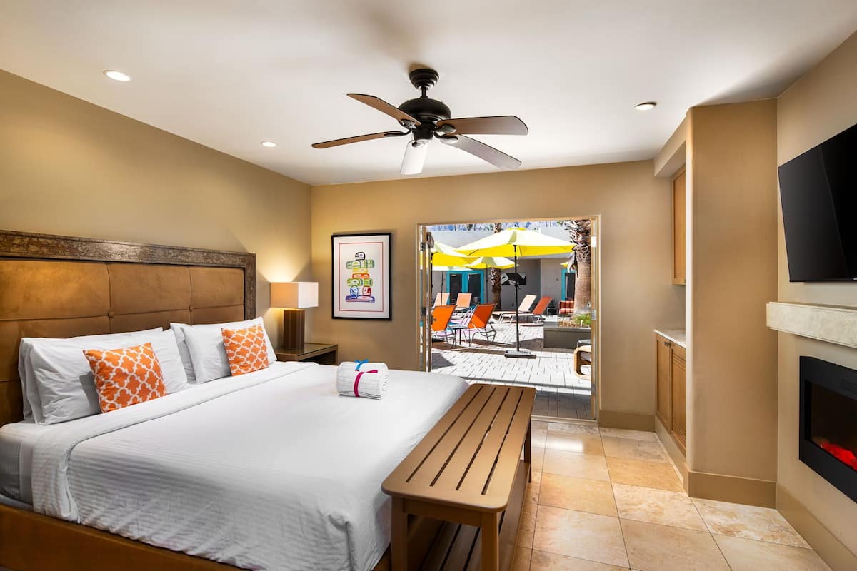 A hotel room with a bed, a ceiling fan, a TV, and a view of an outdoor seating area with umbrellas.