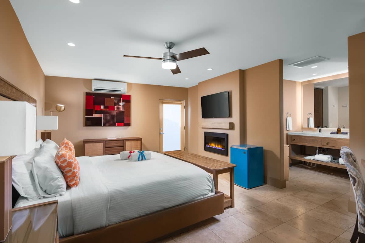 A modern hotel room with a bed, a desk, a television, a fireplace, and a bathroom with a mirror and sink.