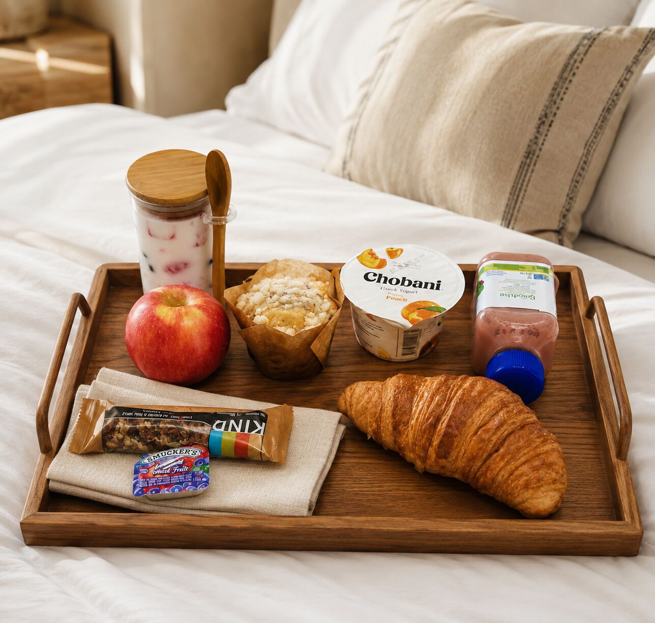 A breakfast tray with a croissant, apple, yogurt, and a bottle of juice on a bed.