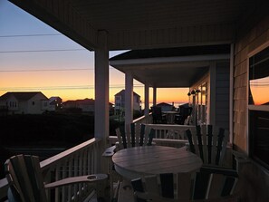 En træterrasse med et rundt bord og stole, med udsigt til en solnedgang og huse i baggrunden.