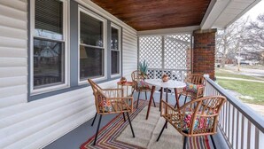 A covered porch with a wooden ceiling, white siding, and a lattice screen. There are wicker chairs and a small round table with a white tablecloth.