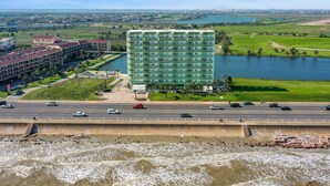 A multi-story hotel with a green facade, located near a body of water and a highway. - Pelicondo | Ocean Front Condo with Beach View (Galveston)