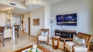 A spacious living area with a large flat-screen TV mounted on the wall, a dining area with bar stools, and a cozy seating arrangement with two armchairs and a small round table. - Pelicondo | Ocean Front Condo with Beach View (Galveston)