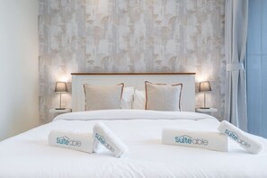 A neatly made bed with white linens and pillows, two bedside lamps, and a patterned wallpaper background.