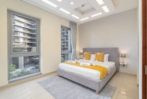 A modern bedroom with a large window, a bed with a gray and yellow bedspread, and a patterned rug. - Sophisticated and Elegant 2BR in Downntown Dubai (Dubai)