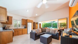 A spacious living area with a dark-colored sofa and ottoman, wooden cabinets, a kitchen with a refrigerator, and a large window offering a view of greenery. - Casa linda Villa 918 (Sosúa)