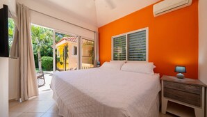 A bedroom with a large bed, an air conditioner, a nightstand with a lamp, and a view of a garden through a sliding glass door. - Casa linda Villa 918 (Sosúa)